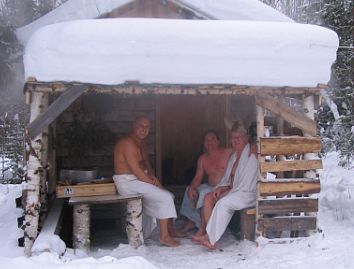 Three men in towels sitting outside a wooden shack.